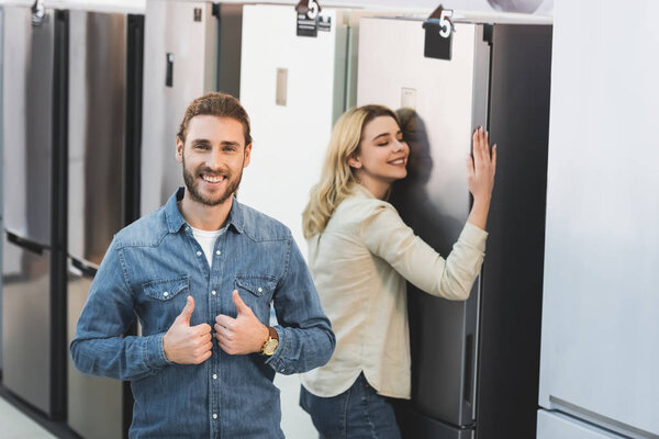 избирательный фокус улыбающегося парня, показывающего симпатии и девушки, касающихся фона в магазине бытовой техники
 