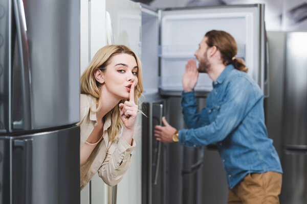 selective focus of girlfriend showing shh and boyfriend looking at fridge on background in home appliance store 