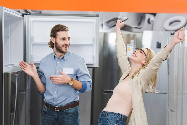 consultant pointing with hands at fridge and smiling woman showing yes gesture in home appliance store