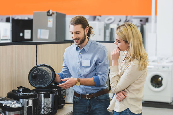 smiling consultant pointing with hand at slow cooker and pensive woman looking at it in home appliance store 