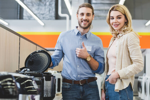 smiling consultant showing like near slow cooker and woman looking at camera in home appliance store 