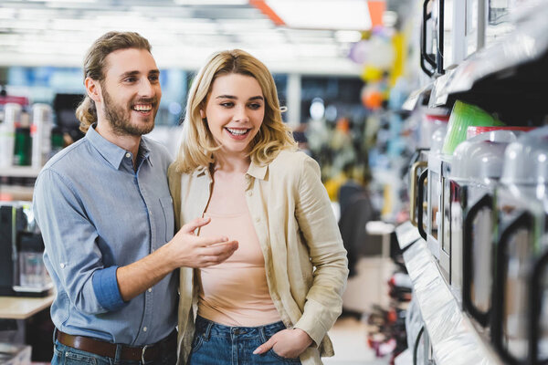 smiling consultant pointing with finger at microwave and talking with woman in home appliance store 
