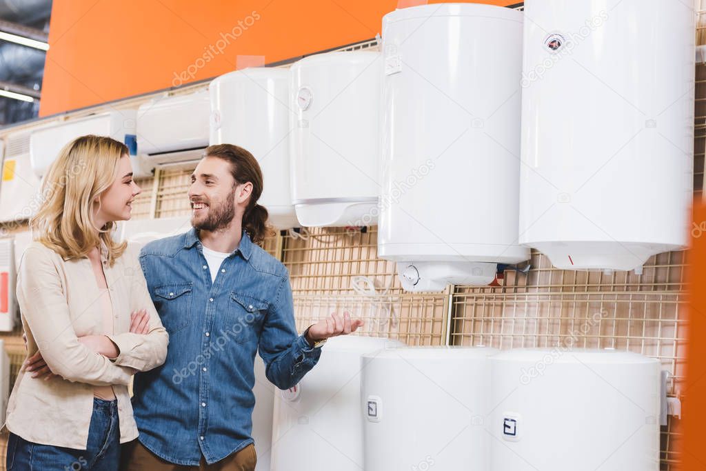 smiling boyfriend and girlfriend talking near boilers in home appliance store 