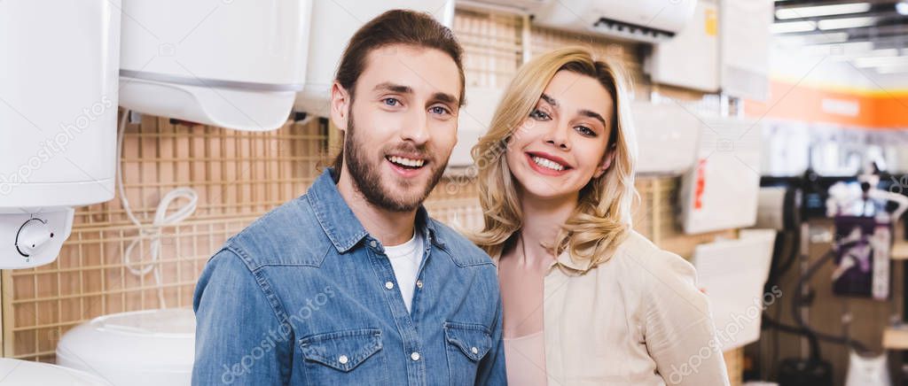 panoramic shot of smiling boyfriend and girlfriend standing near boilers in home appliance store 