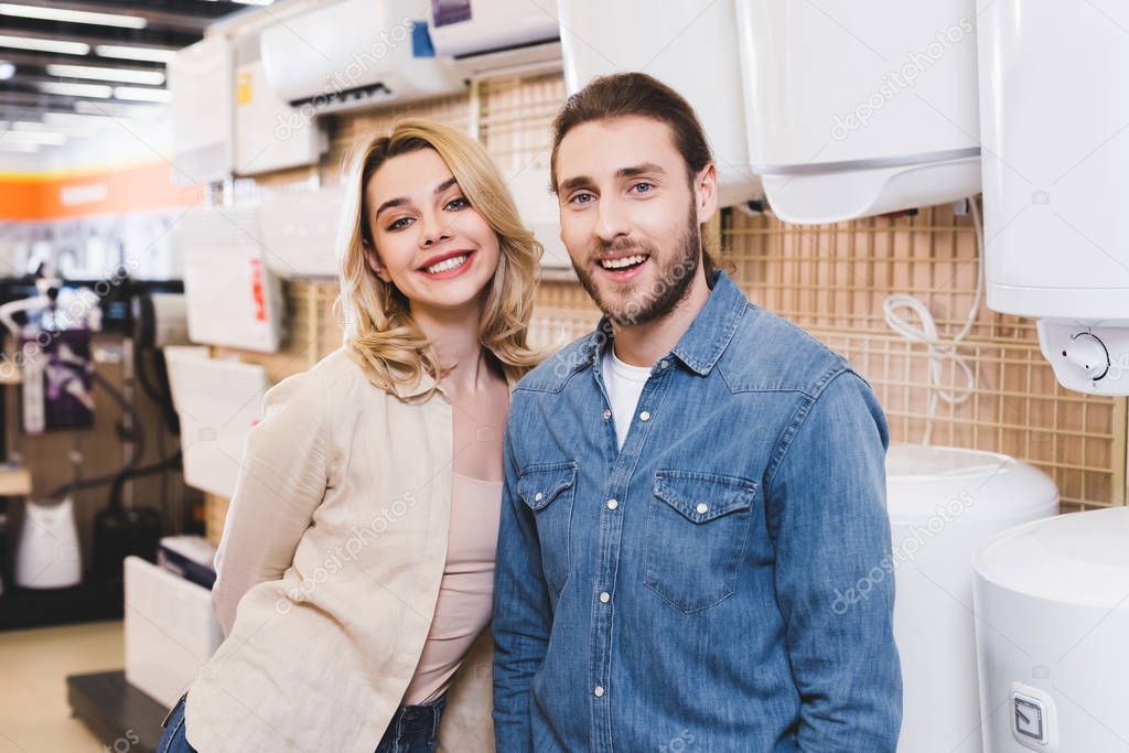 smiling boyfriend and girlfriend standing near boilers in home appliance store 