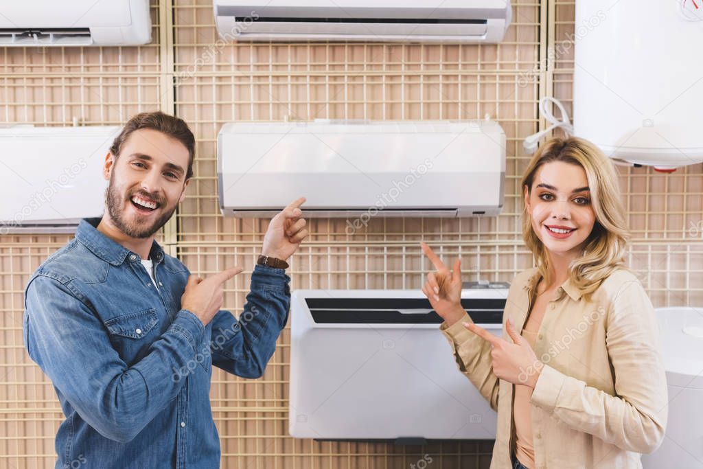 Smiling boyfriend and girlfriend pointing with fingers at air conditioner in home appliance store