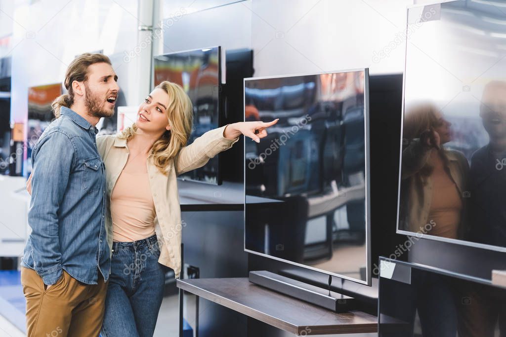 smiling girlfriend pointing with finger at tv and looking at shocked boyfriend in home appliance store 
