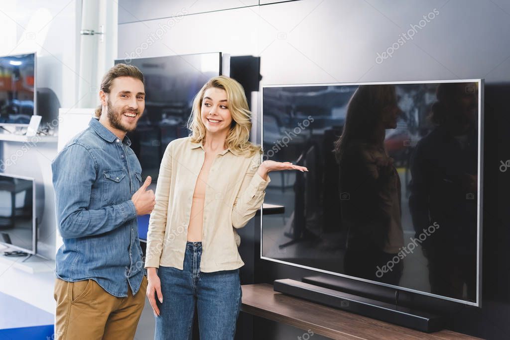 smiling boyfriend showing like and girlfriend pointing with hand at tv in home appliance store 