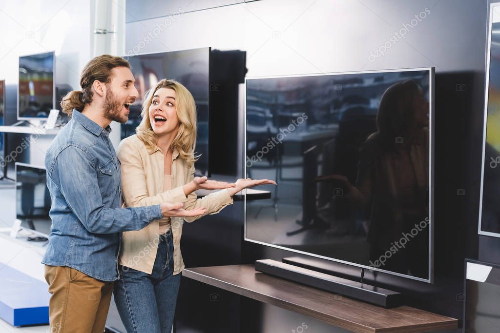 shocked boyfriend and girlfriend pointing with hands at tv in home appliance store 