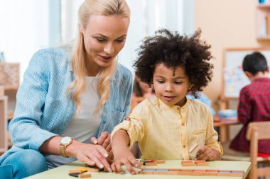 Montessori sınıfında Afrika kökenli Amerikalı bir çocuğa ahşap oyunla yardım eden öğretmen seçici odağı.