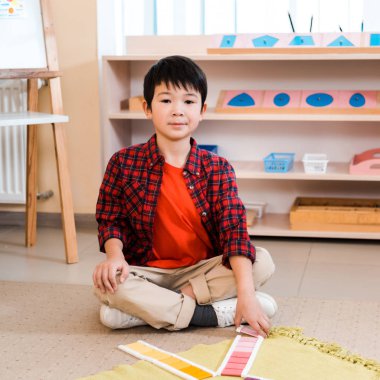 Montessori okulunda yerde renkli oyunlar oynarken Asyalı çocuk kameraya bakıyor.
