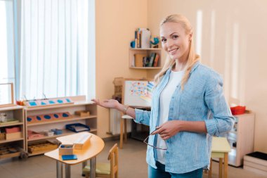 Çekici gülümseme öğretmeni elle işaret ediyor ve Montessori okulunda kameraya bakıyor.