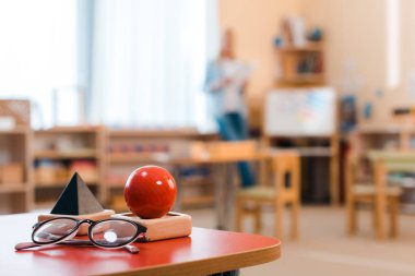Montessori sınıfında arka planda öğretmenle birlikte tahta oyunun ve gözlüklerin seçici odak noktası