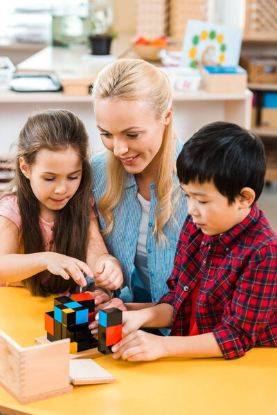 Children playing building blocks by smiling teacher in montessori school