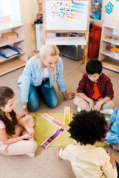 Overhead view of kids and teacher folding colorful game white sitting on floor in montessori school
