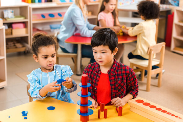 Selective focus of kids folding board game with teacher and children at background in montessori school
