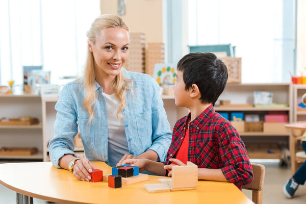 Smiling teacher looking at camera by kid with building blocks in montessori class
