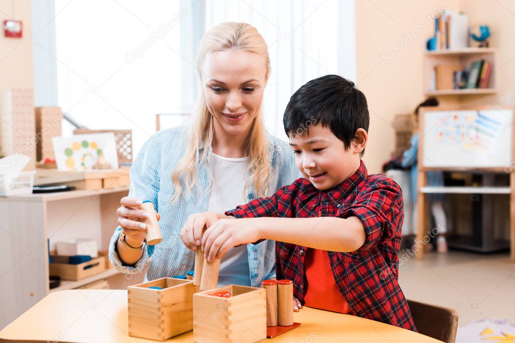 Asian child playing wooden board game by smiling teacher at desk in montessori school