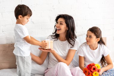 Annesine hediye paketi sunan sevimli çocuk ve anneler gününde elinde çiçek tutan kız kardeş.
