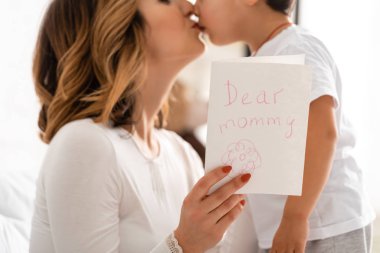 cropped view of happy mother kissing adorable son while holding mothers day card with dear mommy inscription clipart