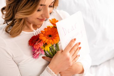 Bir buket gerbera ve anneler günü kartı tutarken gülümseyen mutlu kadın.