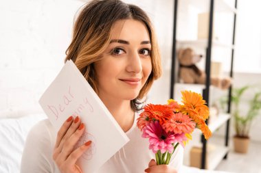 Bir buket Gerbera ve anneler günü kartı tutarken kameraya gülümseyen güzel bir kadın.