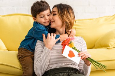 Annesini kucaklayan sevimli çocuk elinde çiçekler ve üzerinde kalp sembolü olan anneler günü kartı tutuyor.