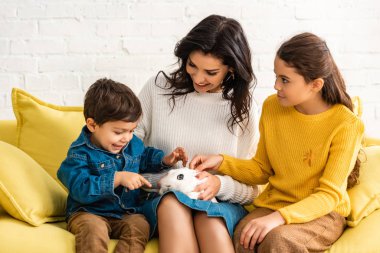 Neşeli anne ve çocuklar sarı kanepede beyaz tavşanla oturuyor.