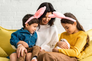 happy woman and cheerful children in bunny ears sitting on yellow sofa with white funny rabbit clipart
