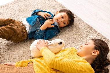 Kız ve erkek kardeşin beyaz komik tavşanın yanında yerde yatarken yüksek açılı görüntüsü.