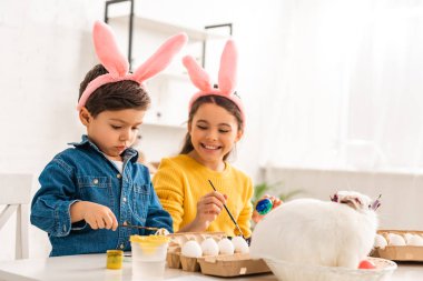 Tavşan kulaklı neşeli çocuklar paskalya yumurtalarını boyarken hasır şapkalı beyaz tavşanın yanında oturuyorlar.