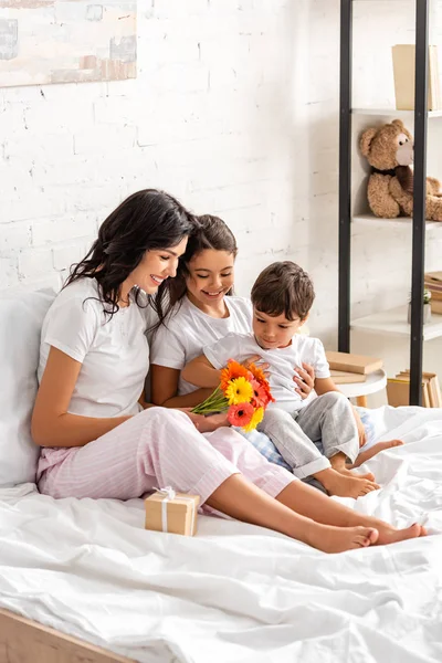 Lycklig Kvinna Hålla Blommor Medan Sitter Sängen Nära Bedårande Barn — Stockfoto