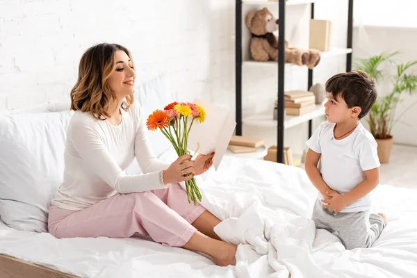 可愛い息子のそばのベッドに座りながら花を手に母の日カードを見る幸せな女性 — ストック写真