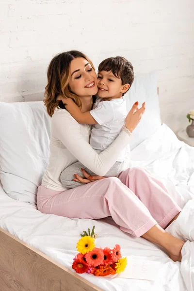在母亲节 快乐的女人坐在花边的床上抱着可爱的儿子 — 图库照片