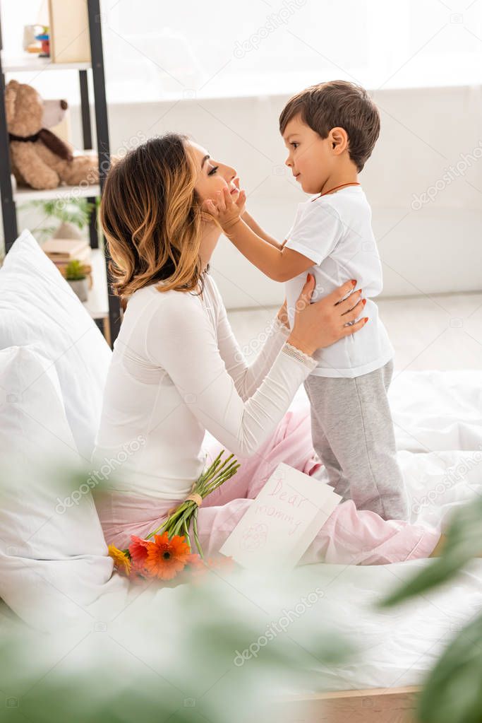 selective focus of cute boy touching face of happy mom sitting in bed near flowers and mothers day card