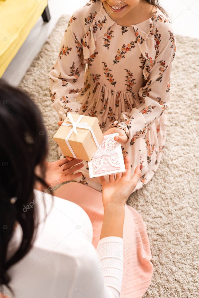 cropped view of elegant child presenting mothers day card and gift box to mom while sitting on floor together