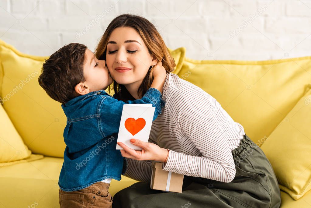 cute boy kissing happy mother holding gift box and mothers day card with heart symbol