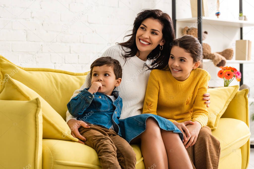 happy mother with son and daughter smiling and looking away while sitting on yellow sofa on mothers day