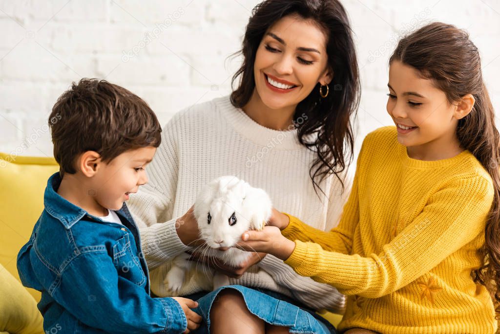 happy children and cheerful mother sitting on sofa with white funny rabbit