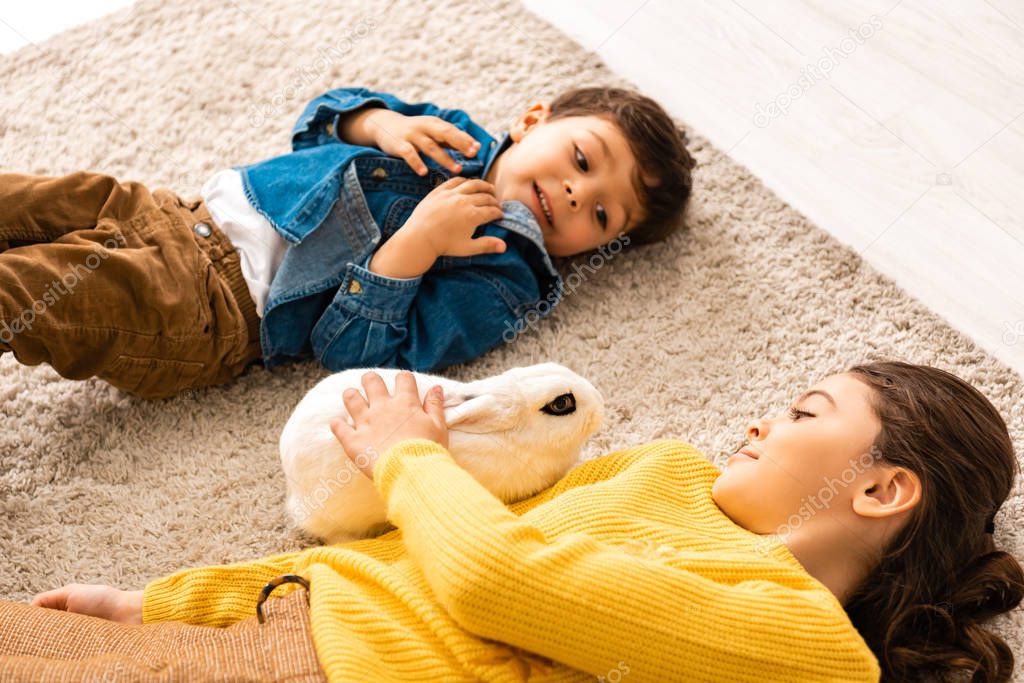 high angle view of brother and sister lying on floor near white funny rabbit