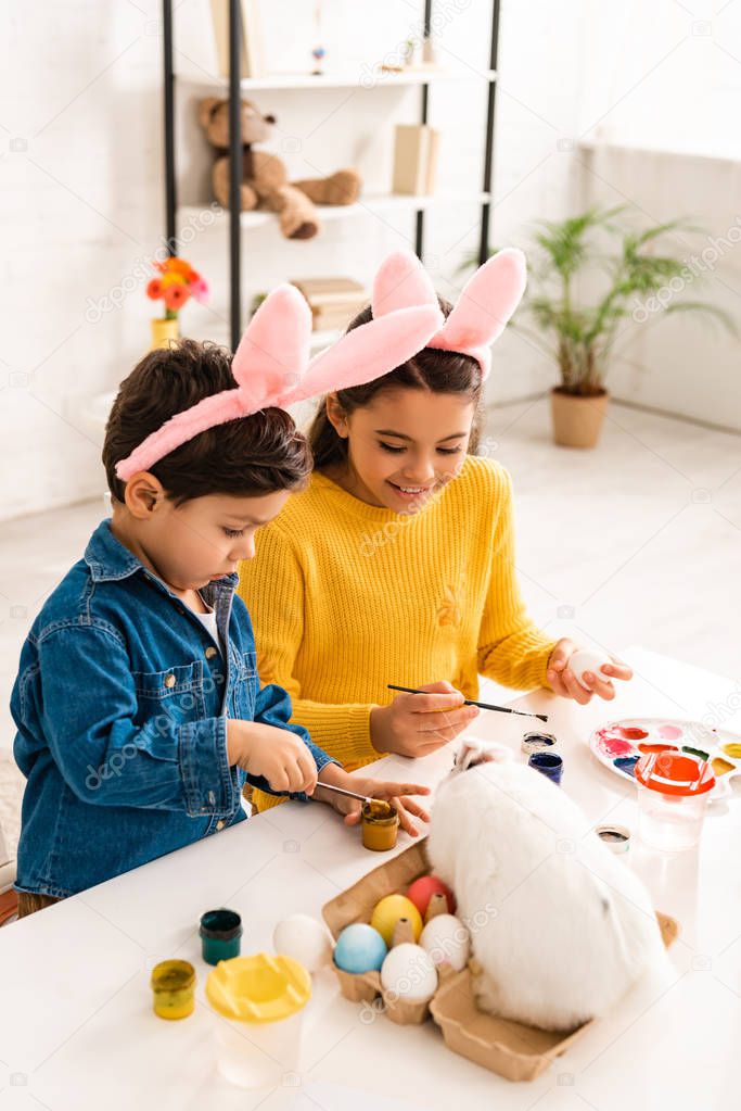 cute brother and sister in bunny ears painting Easter eggs while sitting at desk near white rabbit 