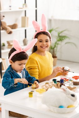 Hasırlı beyaz tavşanın yanında oturmuş kameraya gülümseyen ve Paskalya yumurtalarını tatlı kardeşinle boyayan neşeli bir çocuk.
