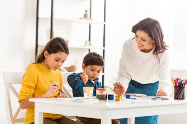 Mutlu kadın masanın yanında duruyor ve çocuklarla birlikte resim çiziyor.