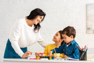 Mutlu anne, masada oturan ve boyalarla resim çizen çocuklara bakıyor.