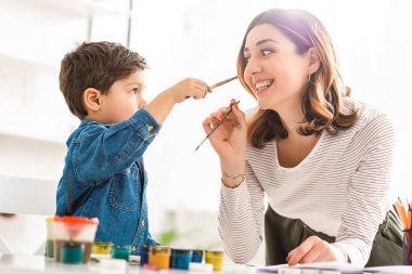 Mutlu anne ve oğul boya kutularının yanında boya fırçalarıyla eğleniyorlar.