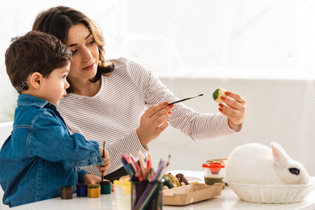 attentive mother and son painting Easter eggs while sitting at table near bunny in wicker