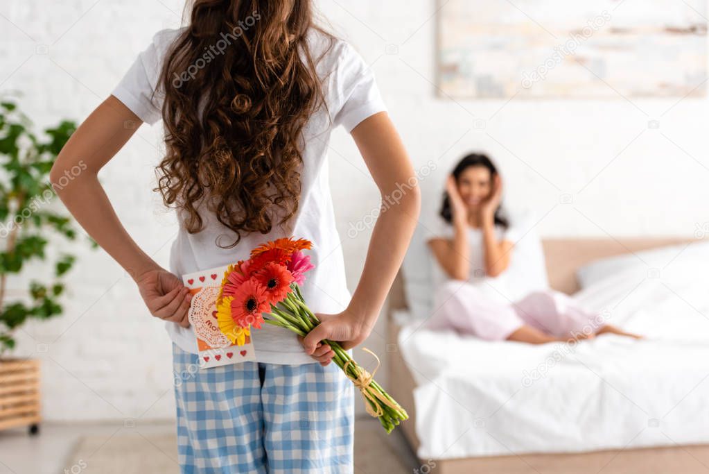 back view of child holding flowers and mothers day card while mom sitting in bed 