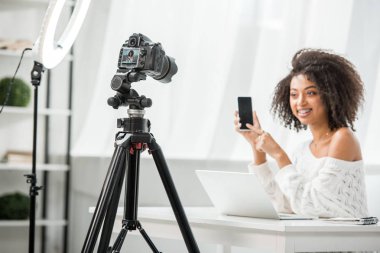 Çekici Afro-Amerikan video blogcusu yakınındaki dijital kameranın seçici odak noktası boş ekranlı akıllı telefonu işaret eden diş telleri. 