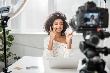 Mutlu Afro-Amerikan video blogcusunun seçici odak noktası. Diş teli takmış dijital kamera yanında kozmetik krem sürüyor. 