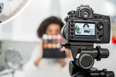 Dijital kameranın seçici odağı kıvırcık Afrika kökenli Amerikalı video blogcusu yüzünü kapatırken göz farı paletini ekranda tutuyor 